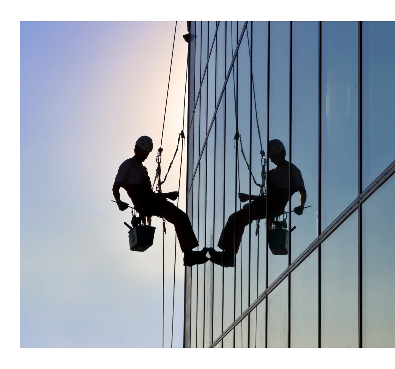 A silhouetted person wearing a helmet is suspended on a harness, showcasing exceptional janitorial services as they clean the windows of a tall glass building at sunset. Their reflection shimmers in the glass.