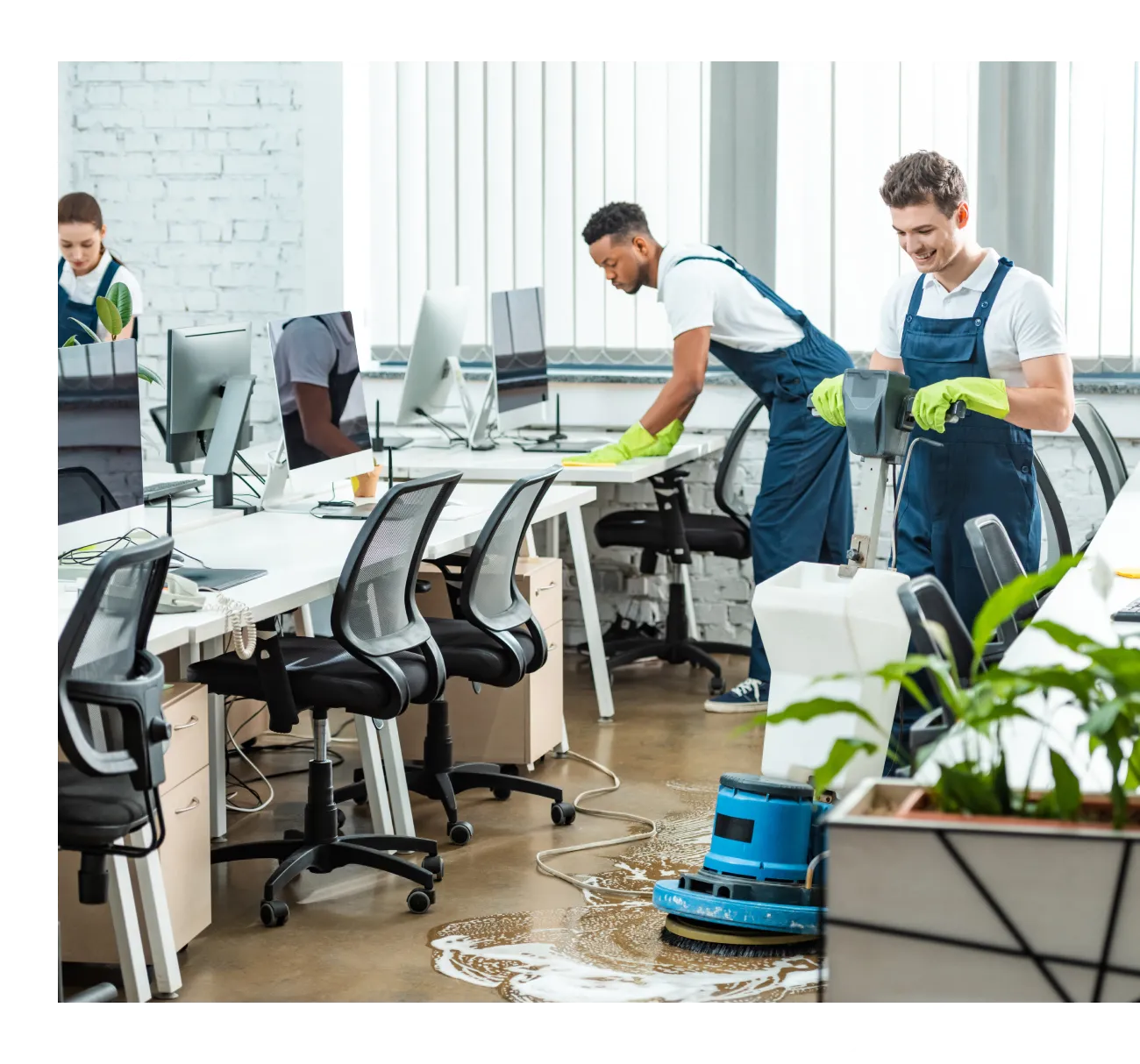 The commercial cleaning crew works diligently in a modern office space, with one person mopping the floor and two others dusting near desks equipped with computers.